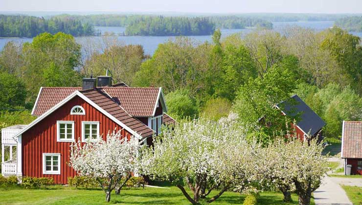 Schwedenrotes Holzhaus mit Blumengarten und Bäumen im Vordergrund und einem See im Hintergrund